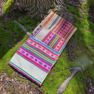 May include: Three woven fabric squares with colorful geometric patterns. The squares are folded on top of each other, with the largest square on the bottom and the smallest square on top. The squares are lying on a bed of green moss.