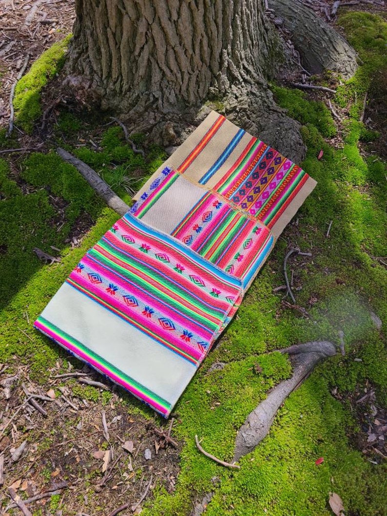 May include: Three woven fabric squares with colorful geometric patterns. The squares are folded on top of each other, with the largest square on the bottom and the smallest square on top. The squares are lying on a bed of green moss.