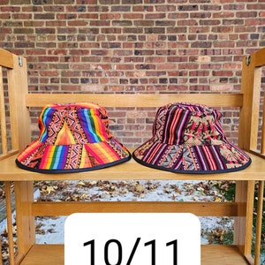 K&ouml;nnte beinhalten: Zwei bunte Bucket-Hats mit geometrischen Mustern. Der Hut links ist orange, gelb, rot und blau. Der Hut rechts ist schwarz, rot und wei&szlig;. Die H&uuml;te befinden sich auf einem Holzregal mit der Nummer 10/11 auf einem wei&szlig;en Etikett.