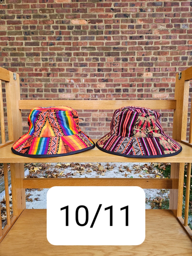 K&ouml;nnte beinhalten: Zwei bunte Bucket-Hats mit geometrischen Mustern. Der Hut links ist orange, gelb, rot und blau. Der Hut rechts ist schwarz, rot und wei&szlig;. Die H&uuml;te befinden sich auf einem Holzregal mit der Nummer 10/11 auf einem wei&szlig;en Etikett.