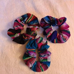 May include: Three colorful hair scrunchies with a woven pattern. The scrunchies are made of fabric and have a variety of colors, including pink, blue, green, and black.