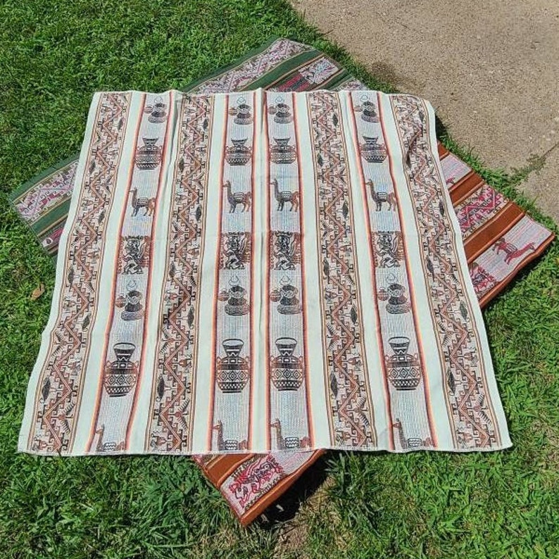 Puede incluir: Un tapiz tejido blanco con un patr&oacute;n repetitivo de llamas, macetas y dise&ntilde;os geom&eacute;tricos. El tapiz tiene un esquema de color marr&oacute;n, rojo y verde.