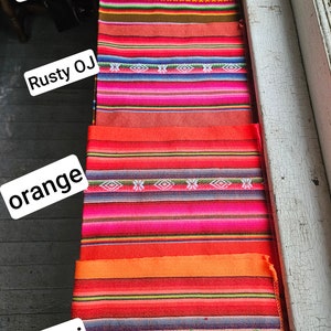 May include: A close-up of five colorful woven fabric samples. The samples are arranged vertically, with the top sample being brown, followed by rusty orange, orange, and neon orange. The bottom sample is a vibrant pink with a blue and green pattern.