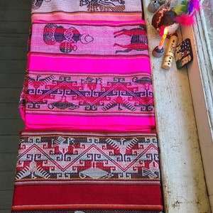 Puede incluir: Una pila de textiles tejidos coloridos con intrincados patrones y dise&ntilde;os. Los textiles son rosas, rojos y blancos con formas geom&eacute;tricas y figuras de animales.