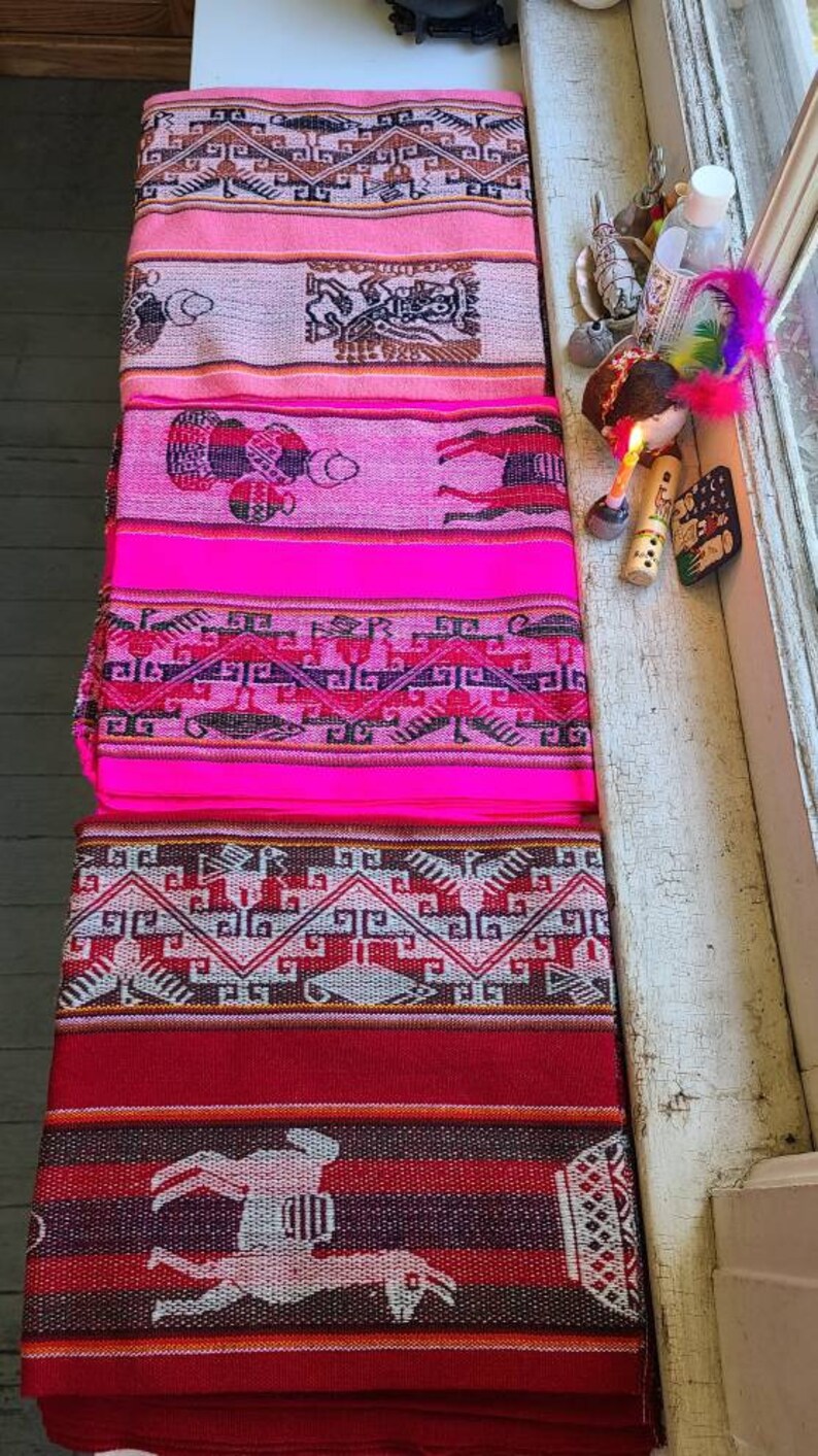 Puede incluir: Una pila de textiles tejidos coloridos con intrincados patrones y dise&ntilde;os. Los textiles son rosas, rojos y blancos con formas geom&eacute;tricas y figuras de animales.
