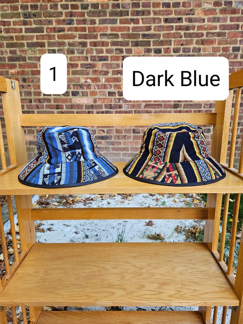 K&ouml;nnte beinhalten: Zwei Bucket-Hats auf einem Holzregal. Der Hut links ist dunkelblau mit einem bunten geometrischen Muster. Der Hut rechts ist schwarz mit einem bunten geometrischen Muster. Der Text "1" und "Dark Blue" ist &uuml;ber den H&uuml;ten sichtbar.