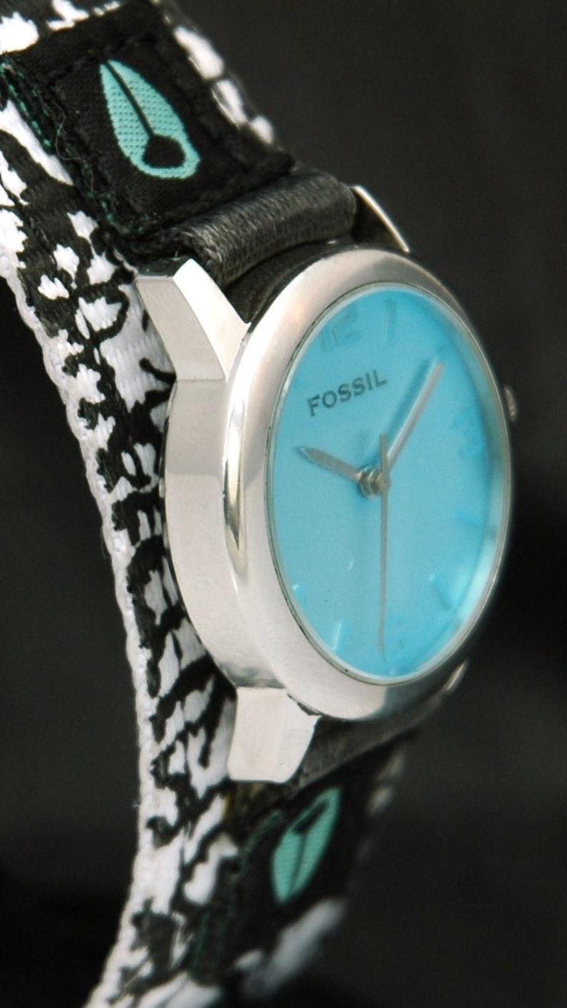 May include: A silver Fossil wristwatch with a blue face and black and white patterned band. The watch face has silver hands and the word "FOSSIL" printed in black.