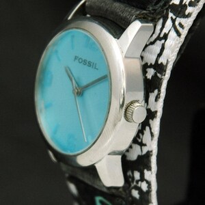 May include: A silver Fossil wristwatch with a blue face and black and white patterned band. The band has a small green logo on it.