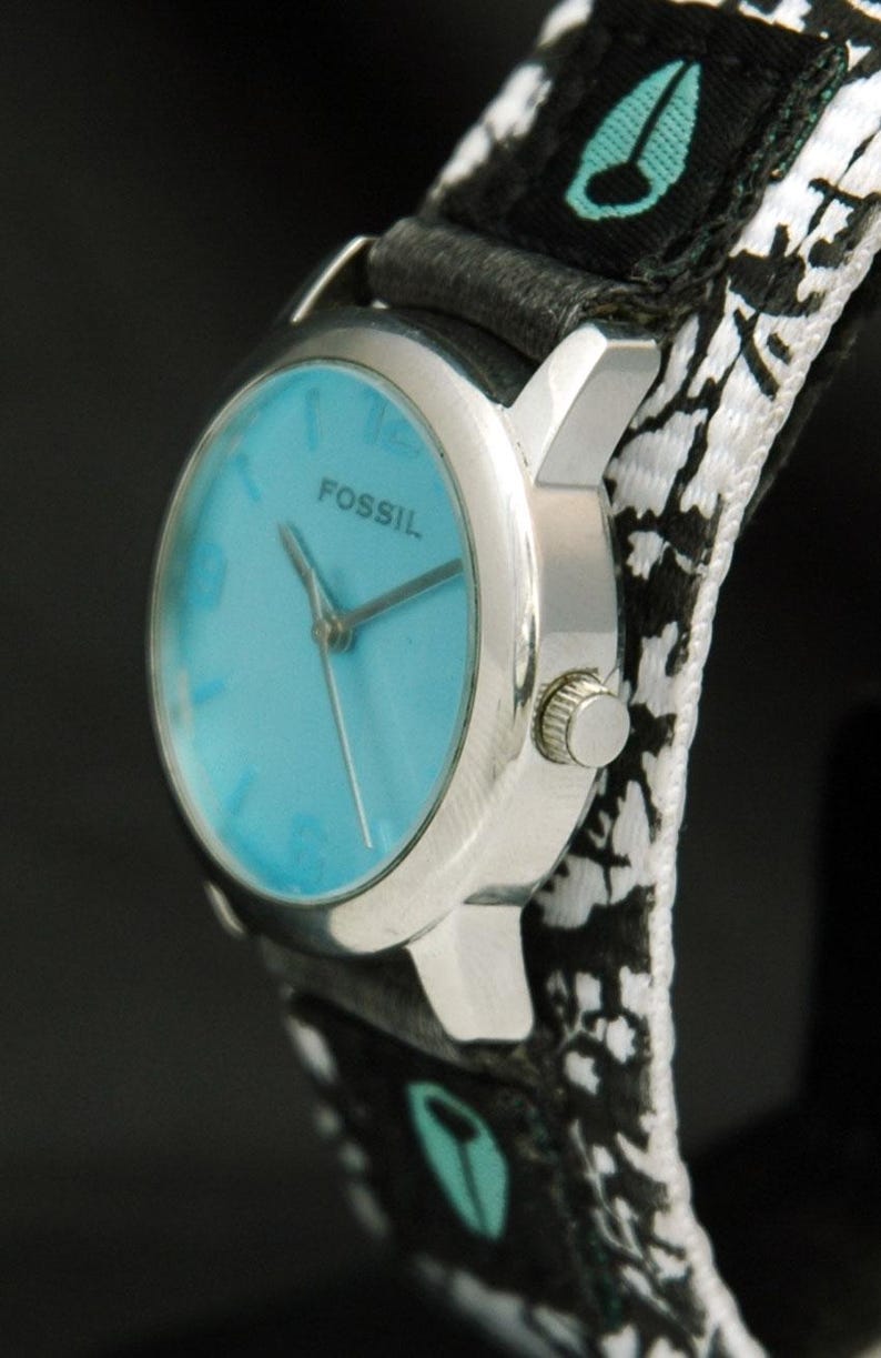 May include: A silver Fossil wristwatch with a blue face and black and white patterned band. The band has a small green logo on it.
