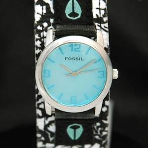 May include: A silver Fossil wristwatch with a light blue face and black and white patterned band. The watch face has black hands and black numbers.