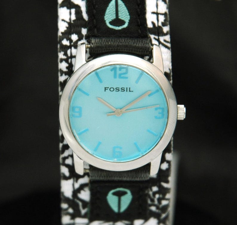 May include: A silver Fossil wristwatch with a light blue face and black and white patterned band. The watch face has black hands and black numbers.
