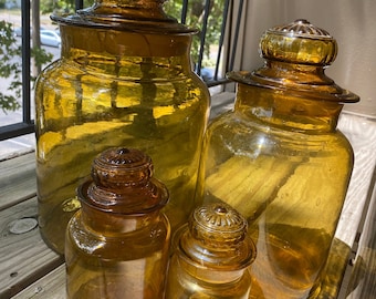 May include: Four amber glass apothecary jars with matching lids. The jars vary in size, with a large jar in the back and smaller jars in the front. The jars are sitting on a wooden surface and are illuminated by sunlight.