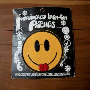 May include: Embroidered iron-on patch featuring a yellow smiley face with black eyes and a red tongue. The patch is on a black background with white text and decorative elements. The text reads "Embroidered Iron-On Patches" and "Cool for Jeans, Tops, Jackets, Hats, Backpacks, etc."
