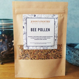 May include: A brown paper bag with a white label that says "JOHN'S PANTRY HERBS & SPICES BEE POLLEN". The label has a floral design and text describing bee pollen. The bag contains yellow and brown bee pollen.
