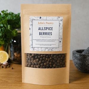 May include: A brown paper bag of allspice berries with a clear window, labeled "John's Pantry." The bag is on a wooden surface with a mortar and pestle, lemon, and parsley in the background. The text on the label reads "Allspice Berries Pimenta Dioica."