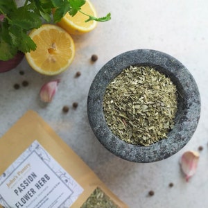 May include: A close-up of a gray stone mortar and pestle with dried passionflower herb inside. The mortar is on a white surface with lemons, garlic, and black peppercorns scattered around it. A brown paper bag with the text "John's Pantry Passion Flower Herb" is in the lower left corner of the image.