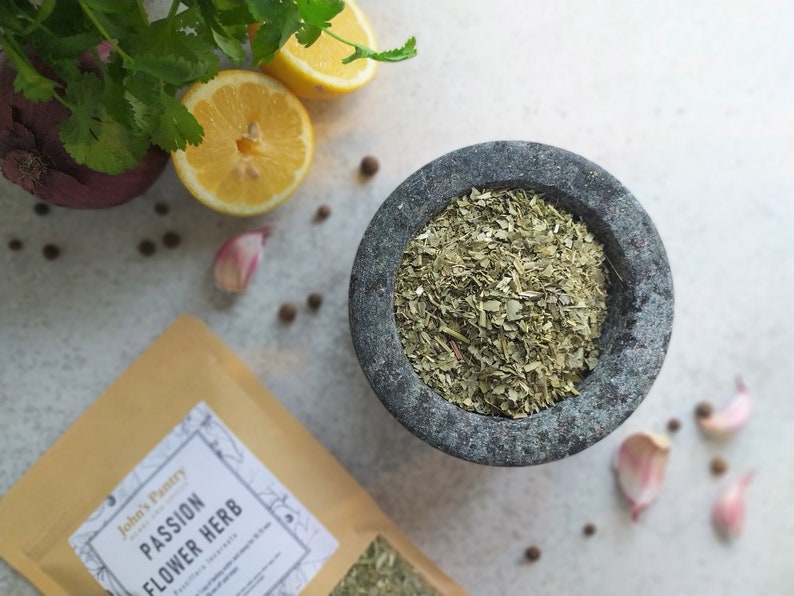 May include: A close-up of a gray stone mortar and pestle with dried passionflower herb inside. The mortar is on a white surface with lemons, garlic, and black peppercorns scattered around it. A brown paper bag with the text "John's Pantry Passion Flower Herb" is in the lower left corner of the image.