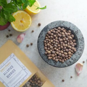 May include: A stone mortar filled with allspice berries, surrounded by fresh ingredients. A package of "John's Pantry Allspice Berries" is visible. Other ingredients include lemon slices, red onion, garlic cloves, and cilantro.