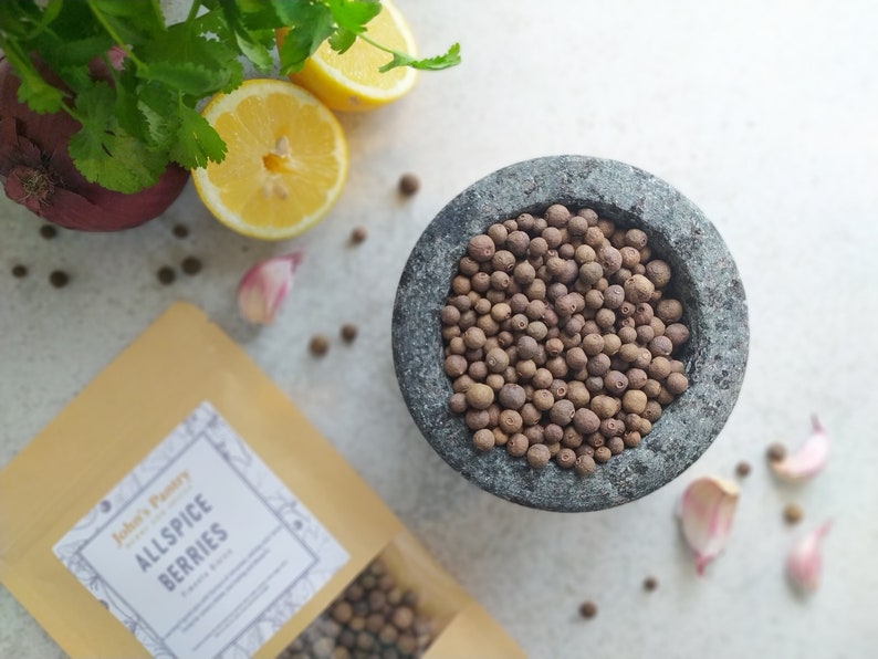 May include: A stone mortar filled with allspice berries, surrounded by fresh ingredients. A package of "John's Pantry Allspice Berries" is visible. Other ingredients include lemon slices, red onion, garlic cloves, and cilantro.
