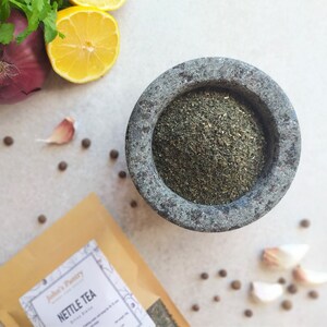 May include: A brown paper bag with the text "Jubilee's Pantry" and "Nettle Tea" printed on it. A mortar and pestle with dried green nettle tea inside. The mortar and pestle are on a white surface with black peppercorns and garlic cloves scattered around.