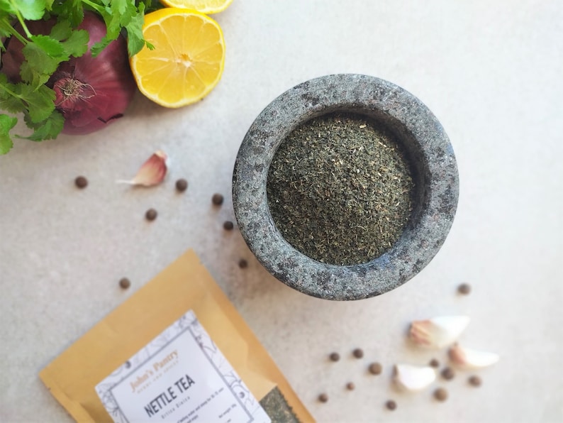 May include: A brown paper bag with the text "Jubilee's Pantry" and "Nettle Tea" printed on it. A mortar and pestle with dried green nettle tea inside. The mortar and pestle are on a white surface with black peppercorns and garlic cloves scattered around.