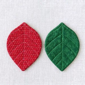 May include: Two leaf-shaped quilted fabric coasters. One is red with a white dotted pattern, the other is green with a snowflake design. Both have stitched vein details and are placed on a white textured surface.
