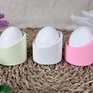 Op de afbeelding: Drie eierrekjes in verschillende kleuren: lichtgroen, wit en roze. Elk rekje heeft een gebogen ontwerp en is gemaakt van een glad materiaal.