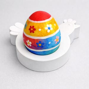 Peut inclure: Un œuf de Pâques coloré avec un dessus rouge, des sections jaunes et bleues, et des motifs floraux. L'œuf repose sur un support blanc en forme de poule, créant un affichage festif et décoratif. Idéal pour Pâques.