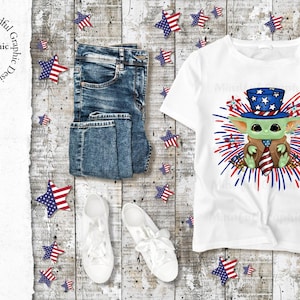 May include: White t-shirt featuring a green character wearing a red, white and blue hat and holding a red, white and blue tie. The character is surrounded by red, white and blue fireworks. The shirt is displayed on a wooden background with blue jeans, white trainers and red, white and blue stars.