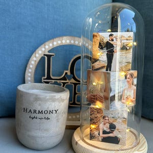 May include: A decorative glass dome with a wooden base, showcasing wedding photos illuminated by fairy lights. A wooden "LOVE" sign and a cement pot with "HARMONY light up life" text are also visible.