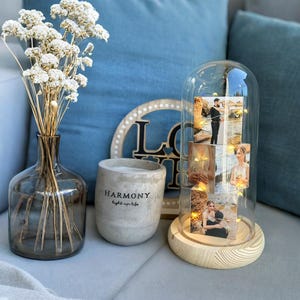 May include: A decorative arrangement featuring a glass cloche with photos illuminated by fairy lights, a vase with dried flowers, a candle holder with the word "HARMONY", and a wooden "LOVE" sign. The items are arranged on a gray surface.