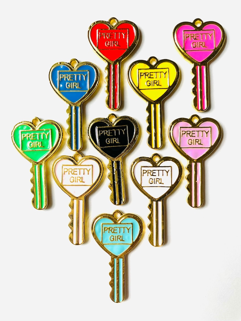 Puede incluir: Diez llaveros de oro con agujeros de llave en forma de coraz&oacute;n. Cada llavero tiene un coraz&oacute;n de esmalte de color diferente con el texto "PRETTY GIRL" en su interior. Los llaveros est&aacute;n dispuestos en c&iacute;rculo sobre un fondo blanco.