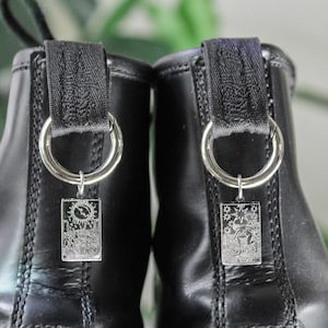 May include: Close-up of two black leather boots with silver charms hanging from the pull tabs. The charms are rectangular with detailed designs. The background is blurred green foliage.