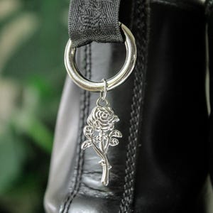 May include: Close-up of black leather boots with silver rose charms hanging from the top rings. The charms are detailed with leaves and a stem. The background is blurred, with a hint of green foliage.