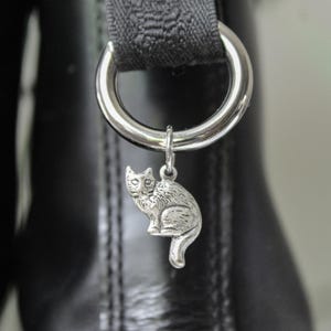 May include: A silver cat charm dangles from a metal ring attached to a black leather item. The charm is a detailed miniature of a sitting cat. The background is blurred, emphasizing the charm and the leather.