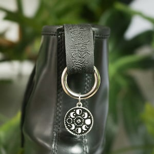 May include: A black leather bag with a silver charm featuring a moon phases design. The charm is attached to a black ribbon loop.