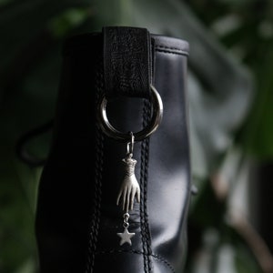 May include: Black leather boot with a silver charm attached to a black strap. The charm is a hand with a star hanging from it.