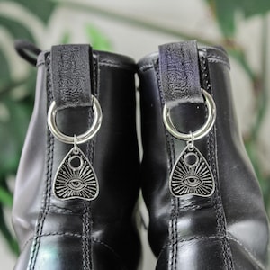 May include: Close-up of two black leather boots with silver-colored Ouija board charms hanging from the back loops. The charms feature an eye design with sun rays. The boots are in focus against a blurred background.