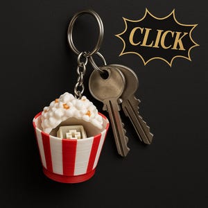 Puede incluir: Un llavero de cubo de palomitas de maíz a rayas rojas y blancas con un tapón de tecla blanco en el interior. El llavero está unido a un llavero plateado con dos llaves plateadas.