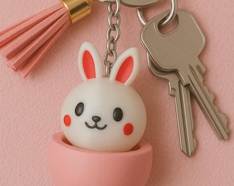 May include: A pink and white bunny keychain with a pink tassel. The bunny is in a pink egg-shaped container. The keychain has a silver key ring and two silver keys.