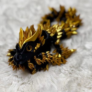 May include: A black and gold dragon figurine with a detailed, spiky design. The dragon's body is black, and the spikes and accents are gold. The figurine is lying on a white, textured surface.