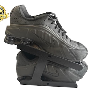 Puede incluir: Zapatilla deportiva negra con el logo de Nike, exhibida en un soporte negro para zapatos. La zapatilla tiene una parte superior texturizada y un diseño de suela único. El soporte es de metal y sujeta el zapato en ángulo.