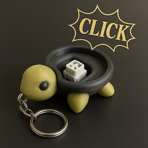 May include: A black and green turtle-shaped keyring with a small, grey, mechanical keyboard switch inside. The keyring has a silver key ring attached to it. The word "CLICK" is written in gold letters on a black starburst.