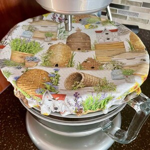 May include: A white stand mixer with a round, white fabric cover featuring a beehive and floral pattern. The cover has a ruffled edge and is secured to the mixer with a clear elastic band.