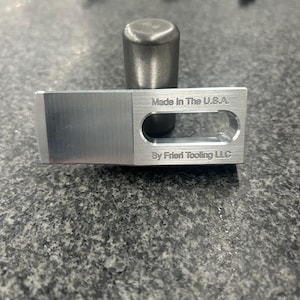 May include: A silver metal tool with a rectangular shape and a hole in the centre. The tool is engraved with the text "Made in the U.S.A. By Frieri Tooling LLC".