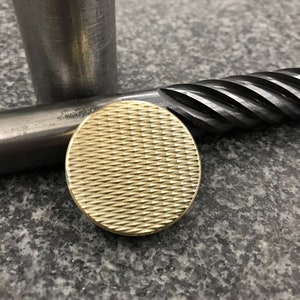 Könnte beinhalten: Ein goldfarbenes, kreisförmiges Metallobjekt mit einem strukturierten, kreuzschraffierten Muster. Das Objekt befindet sich in der Nähe eines zylindrischen Metallstabs und eines schwarzen, spiralförmigen Werkzeugs. Die Gegenstände liegen auf einer grauen, gesprenkelten Oberfläche.