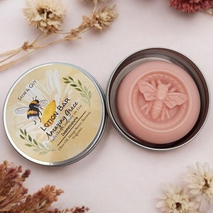 May include: A round tin with a pink lotion bar inside. The lotion bar has a bee design embossed on it. The tin has a label that says "LOTION BAR Amazing Grace" and lists the ingredients as "Olive Oil, Mango Butter, Beeswax, Fragrance".