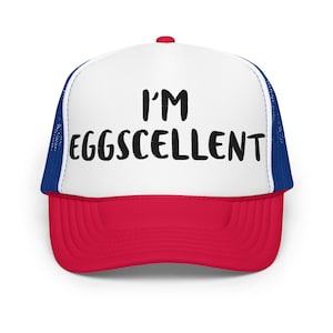 May include: A white and red trucker hat with a blue mesh back. The hat has the text "I'm Eggscellent" in black lettering.