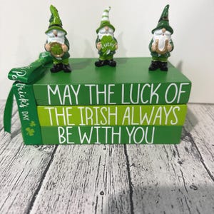 May include: Three green gnomes with shamrocks and a horseshoe stand on top of a stack of three green books. The books are painted with the words "May the luck of the Irish always be with you."