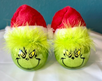 May include: Two green Grinch-themed Christmas ornaments with glitter and a fuzzy green trim. Each ornament is topped with a red Santa hat.